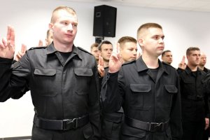 Grupa nowych policjantów ubranych w czarne moro ślubują z uniesionymi prawymi dłońmi z wyprostowanymi dwoma palcami.